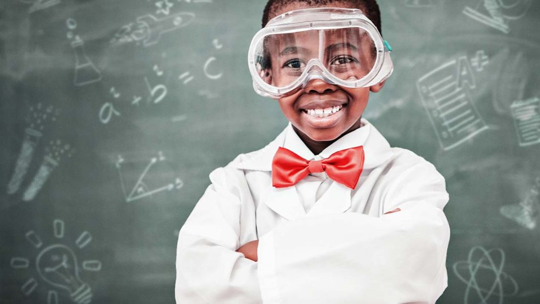 Boy wearing lab coat, red bow tie and safety goggles in a classroom.  Blackboard in the background with different science ico