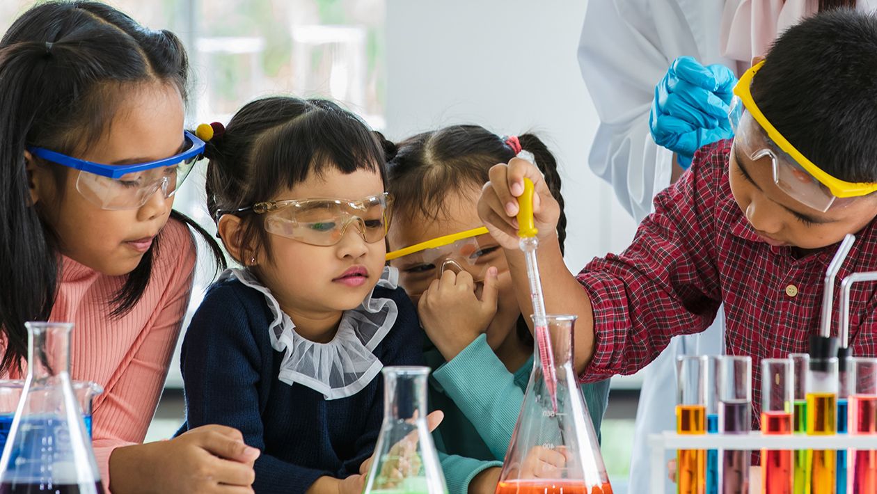 Boy drops something into orange beaker using a pipette in a lab while 3 little girls watch all wearing safety goggles