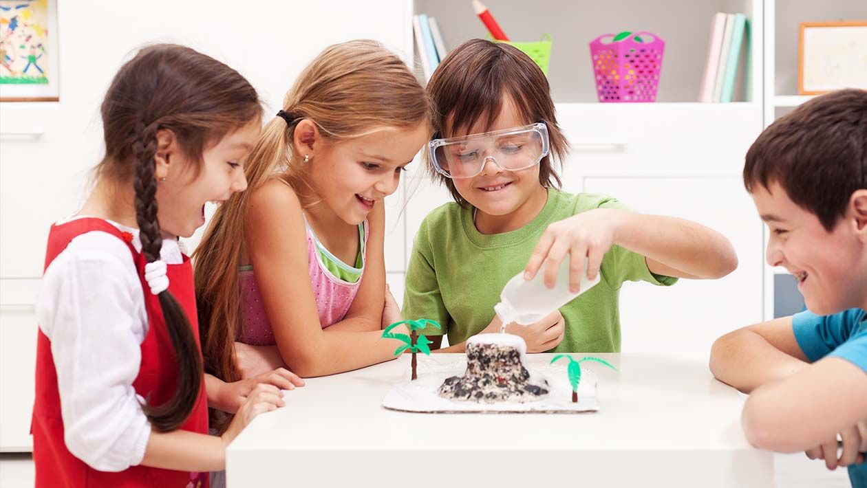 2 boys and 2 girls gathered around a table while one boy is pouring water into a structure to make a volcano