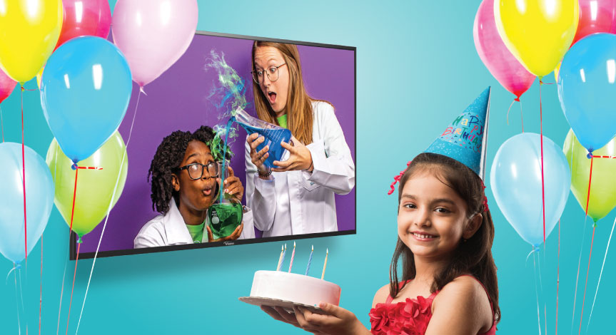 A girl holding a cake wearing a birthday hat watching a tv with a Mad Science instructors pouring a green liquid into a into 